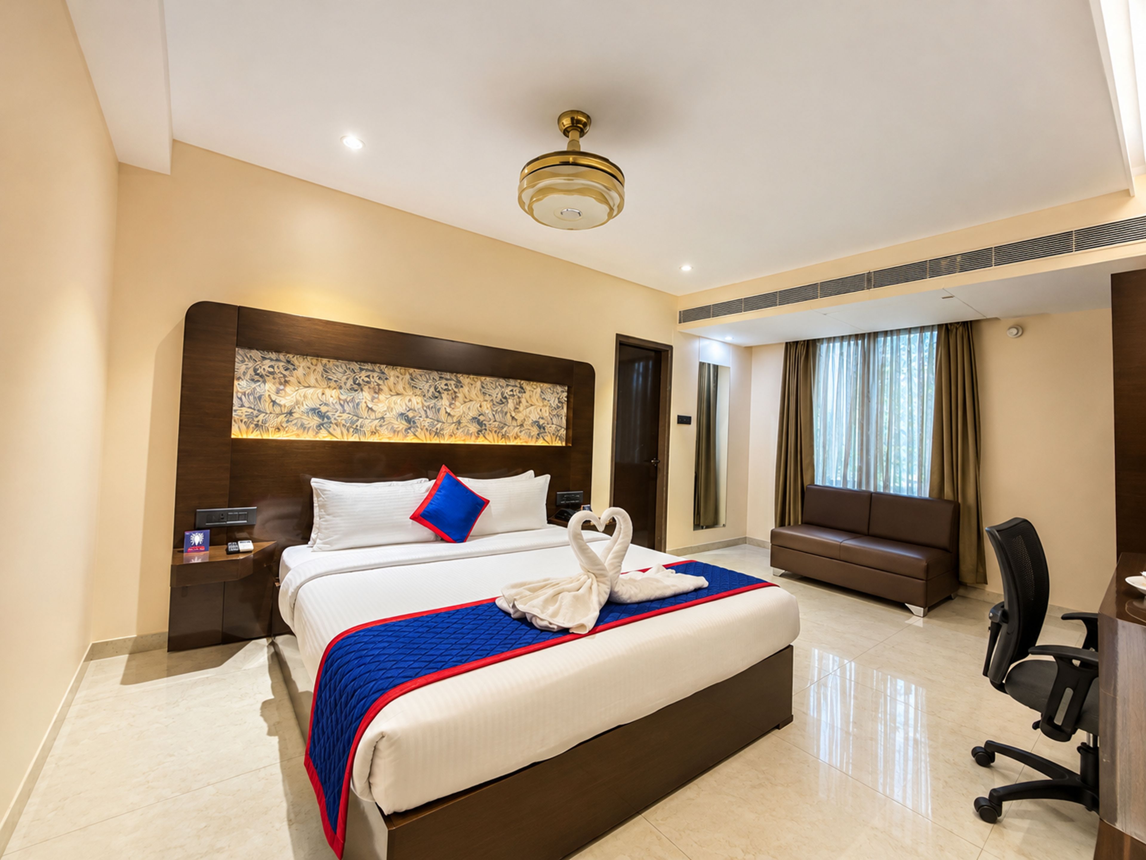 Une chambre d’hôtel avec un grand lit, une tête de lit ornée d’un panneau décoratif, un bureau avec une chaise et une fenêtre avec des rideaux.