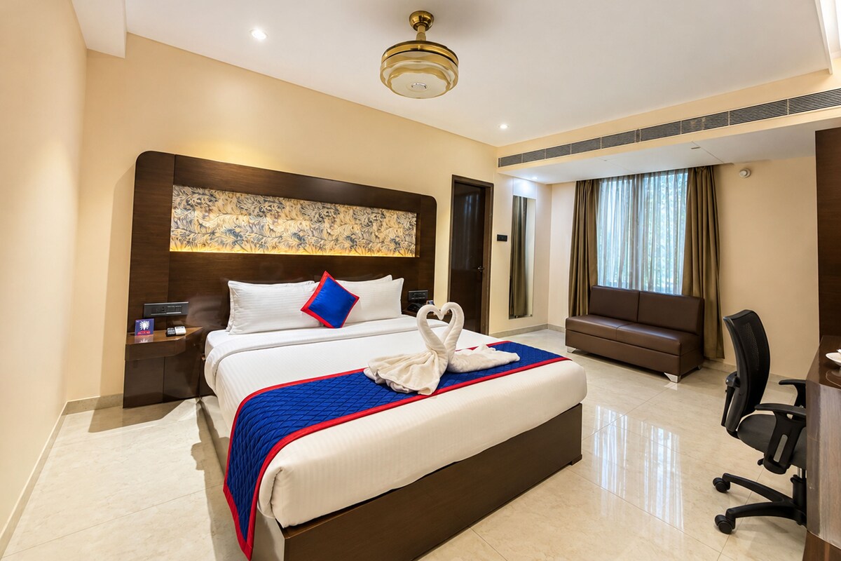 Habitació d'hotel amb un llit gran, un capçal decorat i un escriptori amb cadira, i una finestra amb cortines.