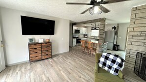 A modern living room with a flat-screen TV, wooden furniture, a ceiling fan, and a kitchen area in the background. - The Hideaway Oliver Springs, Only Min from Windrock Park (Oliver Springs)