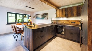 Eine moderne Küche mit Holzausstattung, einer zentralen Kochinsel, Pendelleuchten und Blick ins Freie durch große Fenster.