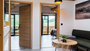 Ein modernes Wohnzimmer mit Holzboden, einem runden Couchtisch mit Pflanze, einem grünen Sofa und einem großen Fenster mit Blick auf eine Terrasse und Grünflächen.