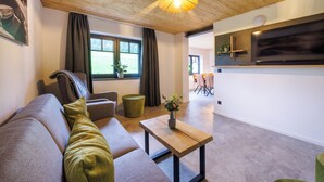Ein modernes Wohnzimmer mit Holzbalkendecke, einem großen Flachbildfernseher, einer Couch, einem Couchtisch und Blick ins Freie.