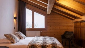 A cozy room with a large bed, a window with curtains, a chair, and a wooden ceiling.