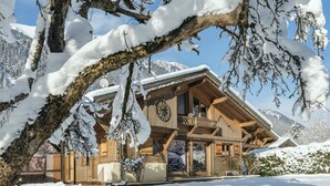 A wooden chalet with a large clock, snow-covered surroundings, and a mountain backdrop.