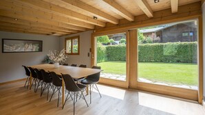 A dining area with a large wooden table and chairs, a framed picture on the wall, and a view of a green garden through large sliding doors.