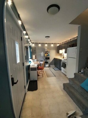 A compact kitchen with a dining area, a small table, and a chair. There is a refrigerator, a stove, and a microwave. The kitchen has a tiled floor and a ceiling light.
