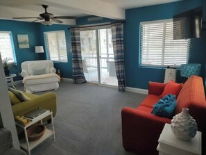 A living room with a ceiling fan, a white sofa, a red sofa, a yellow armchair, and a window with blinds.