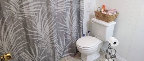 A bathroom with a toilet, a shower curtain, a basket with toiletries, and a roll of toilet paper.