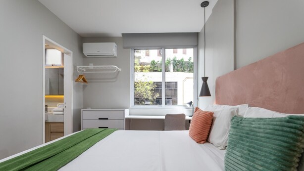 A modern bedroom with a large bed, a desk, and a window with a view of buildings. - Apartment in Copacabana Suite Bi204 (Rio de Janeiro)