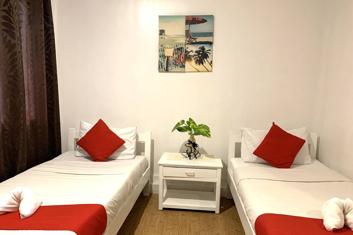 A hotel room with two single beds, each with a red pillow and a white pillow, a small white nightstand with a plant, and a painting of a beach scene on the wall.