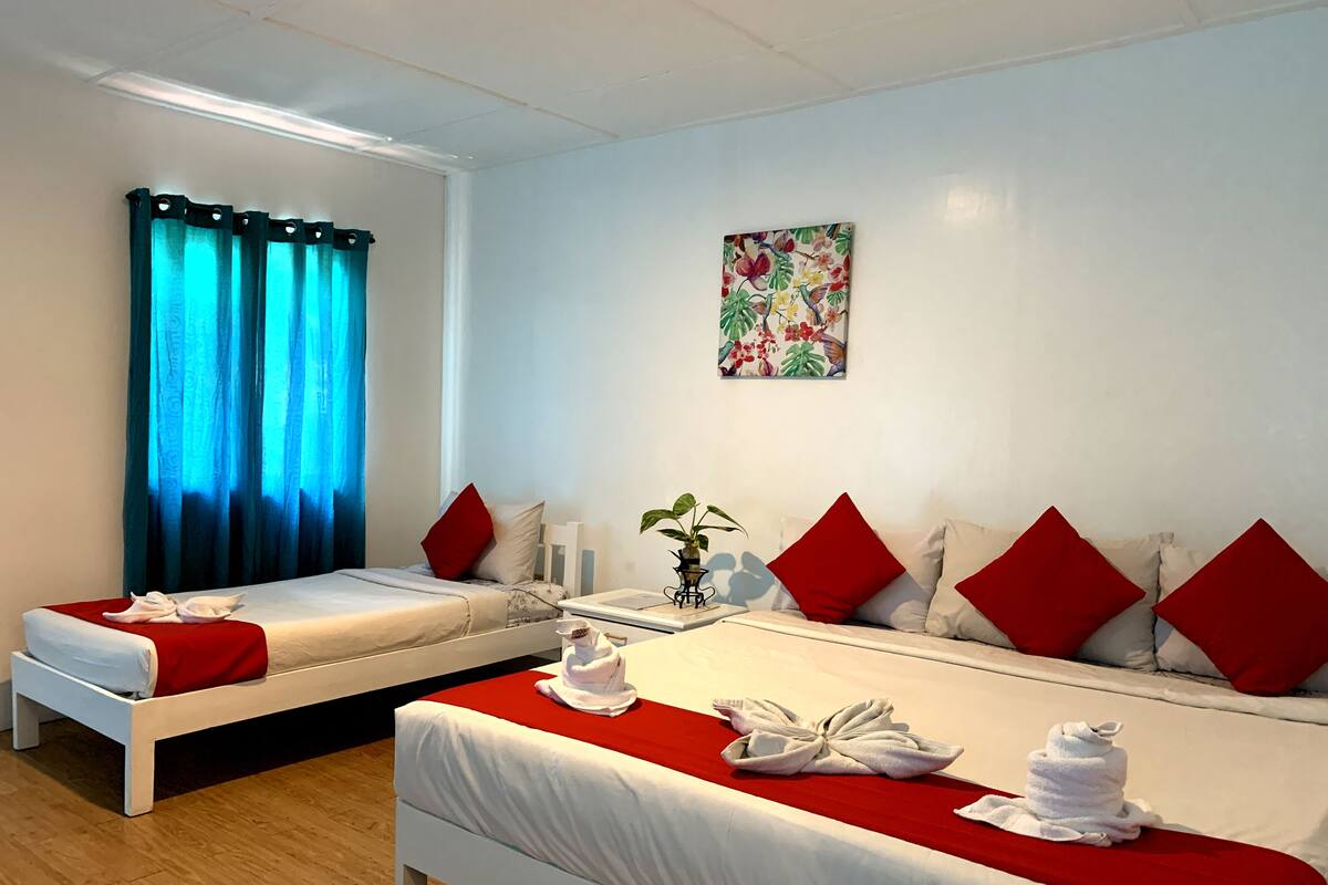 A hotel room with two beds, red and white bedding, a floral painting on the wall, and a window with blue curtains.