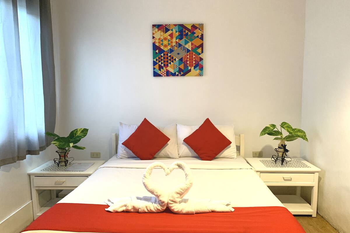 A hotel room with a neatly made bed, two red pillows, a heart-shaped towel, and two potted plants on nightstands.