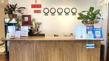 Balcão de recepção com relógios mostrando diferentes cidades, uma cruz e diversos avisos, incluindo opções de pagamento.