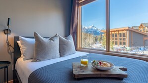 A bedroom with a bed, pillows, a bedside table, a tray with breakfast, a glass of orange juice, and a window with a mountain view. - Scenic Bliss Penthouse | Prime Location (Canmore)