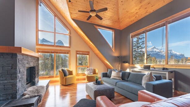 A spacious living room with a stone fireplace, large windows offering mountain views, and a sloped ceiling with a fan. - Scenic Bliss Penthouse | Prime Location (Canmore)