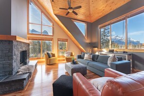 A spacious living room with a stone fireplace, large windows offering mountain views, and a sloped ceiling with a fan. - Scenic Bliss Penthouse | Prime Location (Canmore)