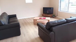 Una sala de estar con dos sofás negros, una mesa de centro de madera, un televisor de pantalla plana sobre un mueble de madera y una ventana con vistas a un edificio.