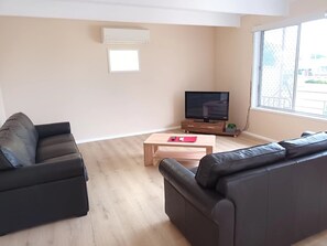 Ein Wohnzimmer mit zwei schwarzen Sofas, einem Holztisch, einem Flachbildfernseher auf einem Holzständer und einem Fenster mit Blick auf ein Gebäude.