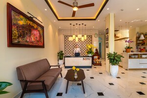 Zona de recepción de un hotel con un sofá, una mesa baja con flores, un mostrador de recepción y un cuadro colgado en la pared.