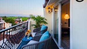 Un balcon avec des fauteuils en osier, une petite table surmontée d’un vase et une plante en pot. Deux lampes murales et une porte-fenêtre coulissante donnent accès à un espace intérieur.