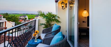 A balcony with wicker chairs, a small table with a vase, and a potted plant. There are two wall-mounted lamps and a sliding glass door leading to an interior space.