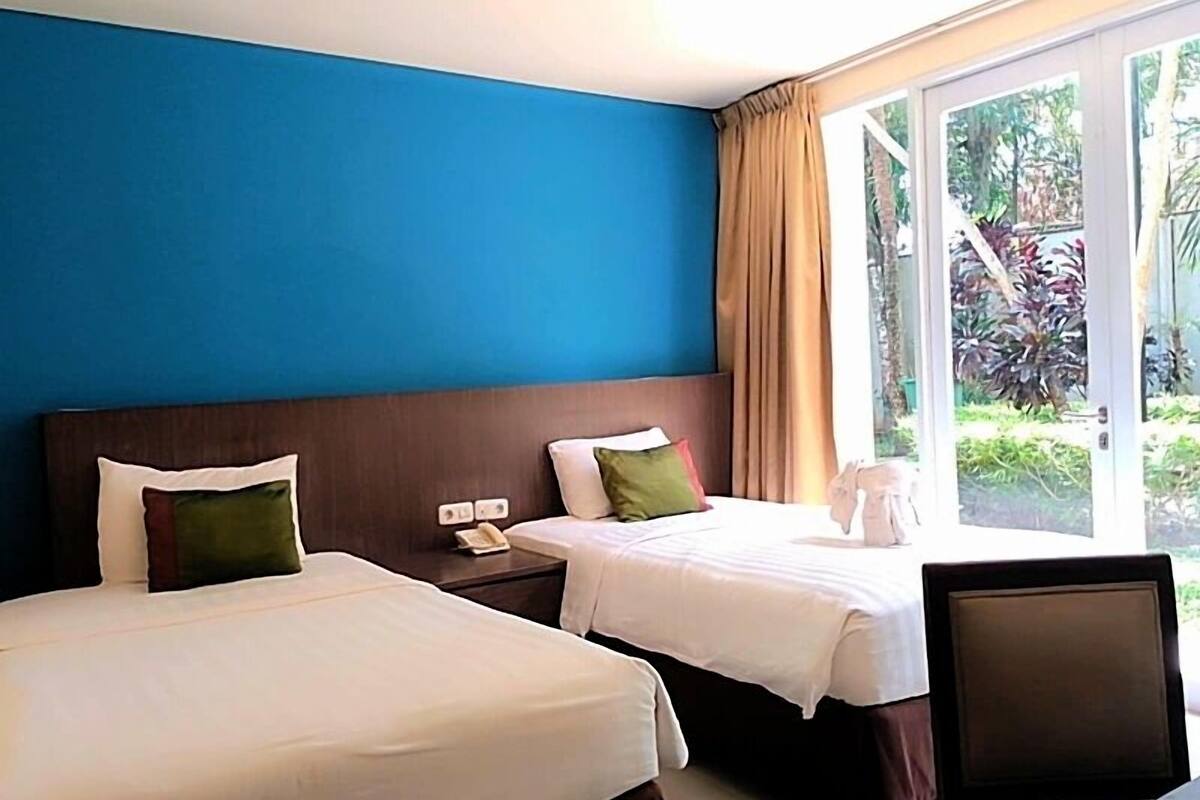 Een hotelkamer met twee bedden, een houten hoofdbord, een klein nachtkastje met een telefoon, en uitzicht op groen buiten het raam.