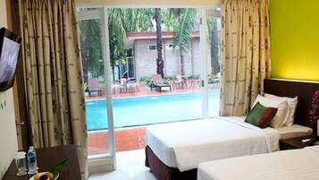 Habitación de hotel con dos camas, televisor, mesa de comedor y una fuente de fruta, con vistas a una piscina y una zona de estar al aire libre.