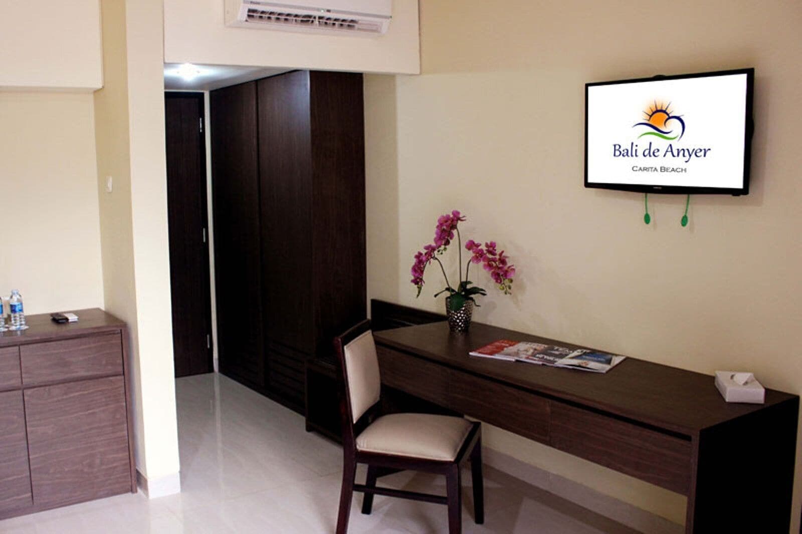 Zona de recepción de un hotel con un mostrador de madera, una silla, un jarrón con flores rosadas y un televisor que muestra el logotipo del hotel.