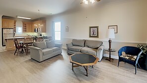 Un salon spacieux avec un canapé gris, une table basse et un fauteuil bleu. On aperçoit au fond une cuisine avec des meubles en bois et un coin repas avec des chaises.