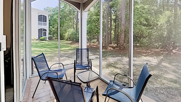 A covered patio with two chairs and a table, overlooking a well-maintained lawn and a white building in the background. - Golf and Beach Getaway Relaxation at its Finest (Pawleys Island)