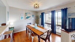 A dining area with a wooden table, white chairs, and a chandelier. There are framed pictures on the wall and a plant near the window. - Heron Marsh Hideaway Split Level Villa (Pawleys Island)