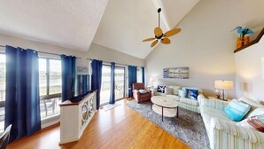 A spacious living room with a wooden floor, a ceiling fan, a television, a sofa set, a coffee table, and a view of the outdoors through large windows with blue curtains. - Heron Marsh Hideaway Split Level Villa (Pawleys Island)