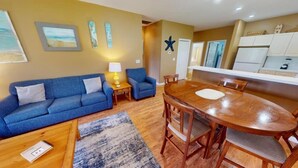 A living room with a blue sofa, a wooden dining table, and a kitchen area with white cabinets.