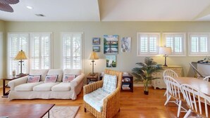 Una sala de estar con sofá, sillón, mesa de comedor y sillas. Hay ventanas con contraventanas, una lámpara y obras de arte en la pared.
