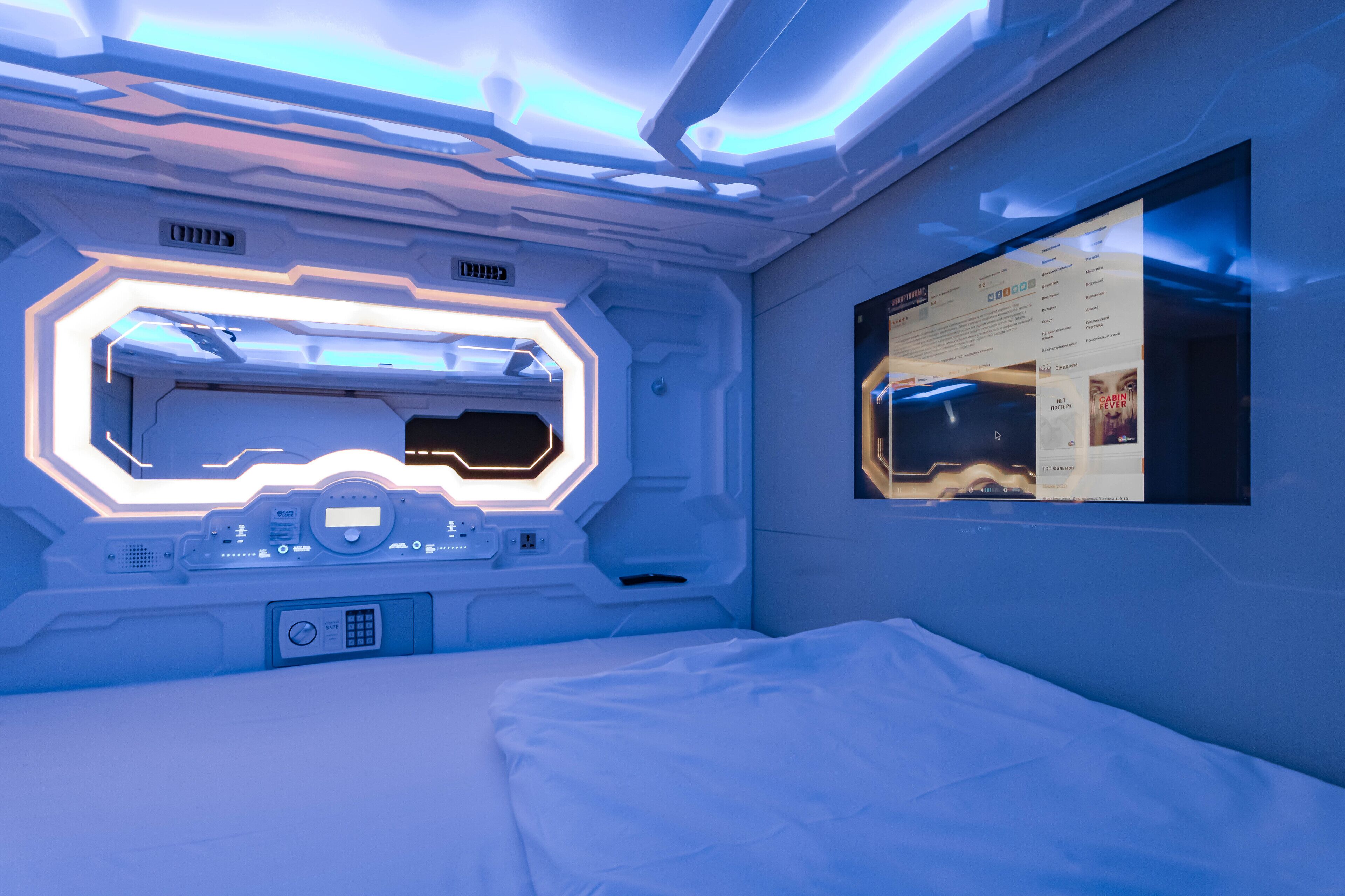 A futuristic room with a central control panel, a bed, and a wall-mounted screen displaying a movie poster.