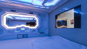 A futuristic room with a central control panel, a bed, and a wall-mounted screen displaying a movie poster.