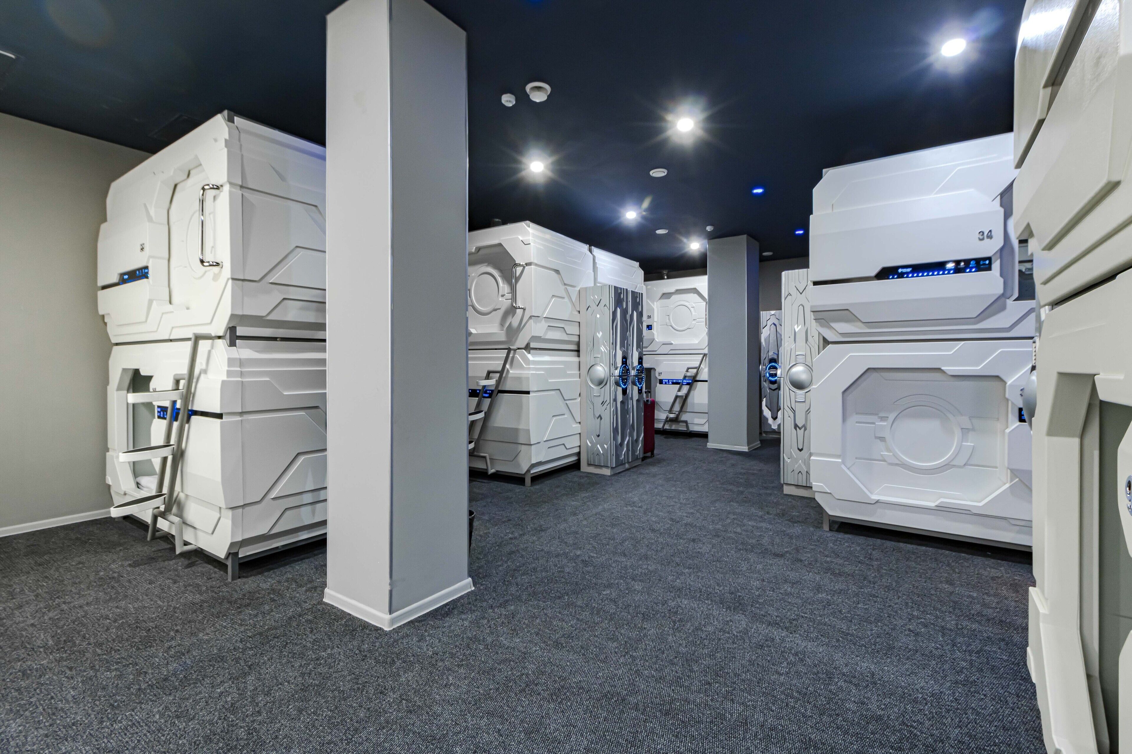 A room with large, white, futuristic-looking machines lined up against the walls, a carpeted floor, and a dark ceiling with recessed lighting.