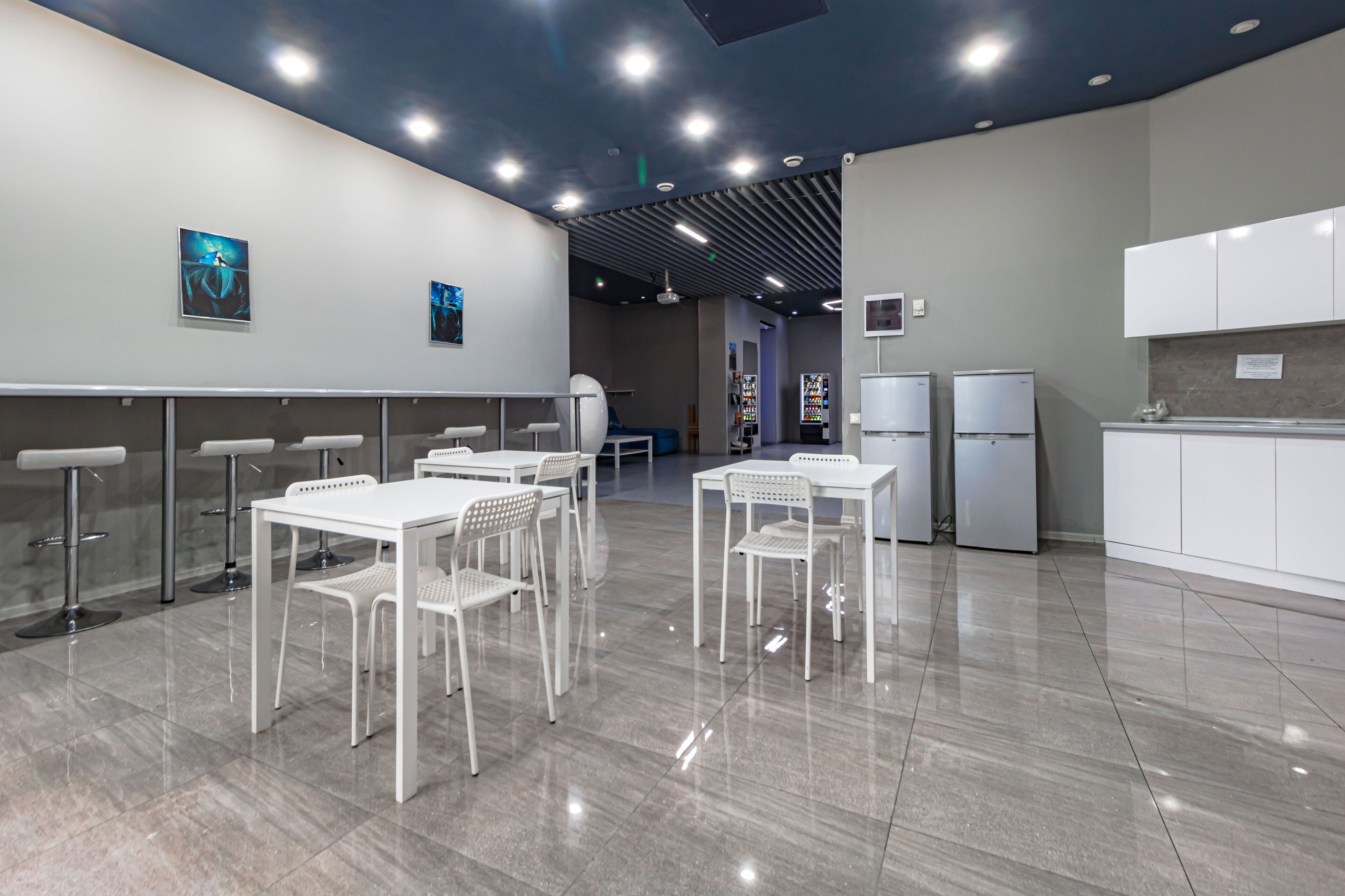 Un comedor moderno y bien iluminado con mesas y sillas blancas, una zona de cocina con armarios blancos e iluminación en el techo.