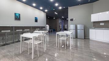 Un comedor moderno y bien iluminado con mesas y sillas blancas, una zona de cocina con armarios blancos e iluminación en el techo.
