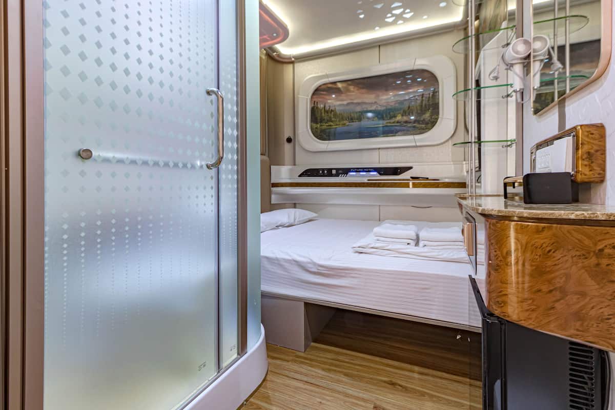Une chambre à coucher compacte et bien éclairée, comprenant un lit, une petite fenêtre offrant une vue agréable, une coiffeuse avec un miroir et un espace douche.