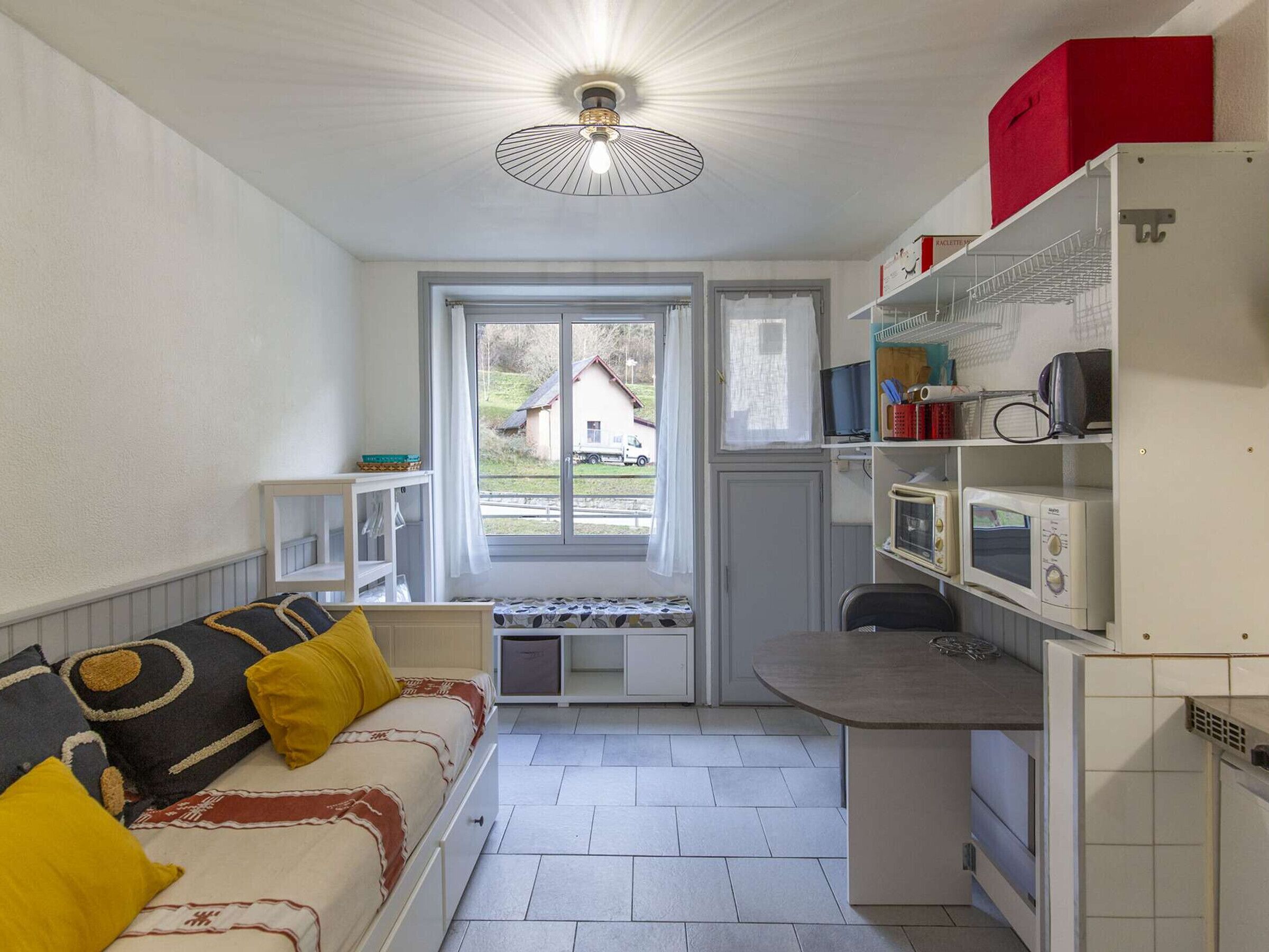 A compact room with a bed, a small kitchenette, and a window with a view of a house and a truck outside.