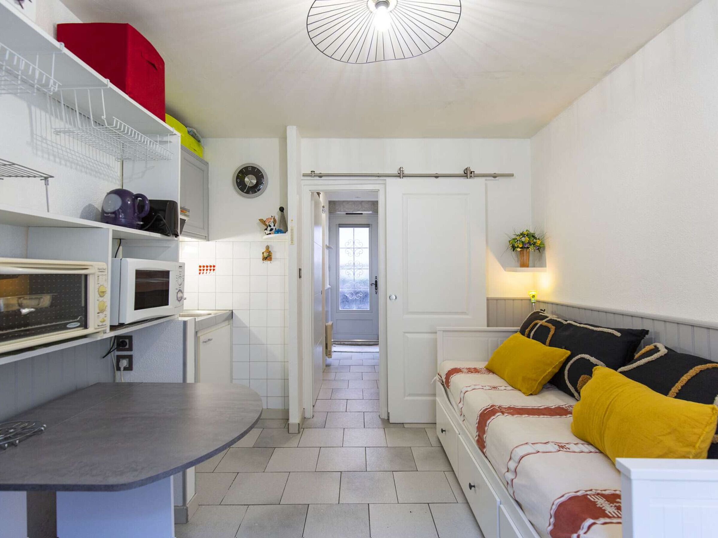A compact kitchen with white appliances, a dining area with a grey table, and a sofa with yellow cushions.