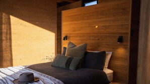 A bedroom with a wooden headboard, a bed with multiple pillows, a bedside table, and a hat on the bed. - Wambal Cabin - luxury in the wilderness (Coxs Creek)