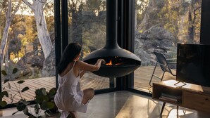 A woman adjusting a modern fireplace in a room with large windows overlooking a forested area. - Wambal Cabin - luxury in the wilderness (Coxs Creek)