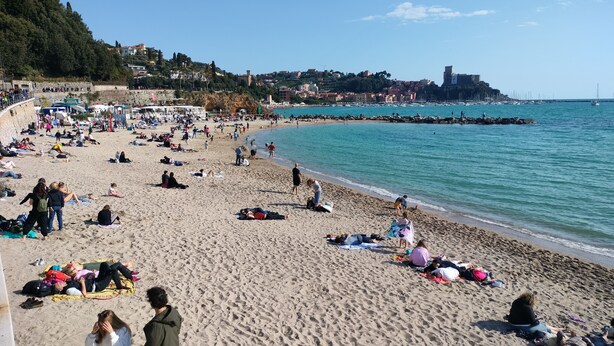 A crowded beach with people sunbathing and swimming, a rocky coastline, and buildings in the background.