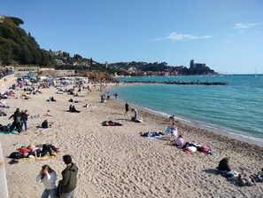 A crowded beach with people sunbathing and swimming, a rocky coastline, and buildings in the background.