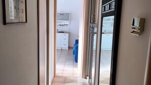 A hallway with a tiled floor, a framed picture on the left, a large mirror on the right, and a doorway leading to a room with a blue chair and a television.