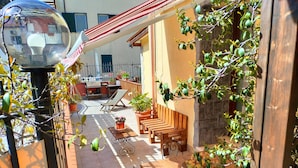 A sunlit patio with a wooden table and chairs, surrounded by potted plants and a red-striped awning.