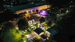 Un espacio para eventos al aire libre con escenario y zona de estar, rodeado de árboles y edificios.