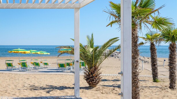 Vista de una playa con muelle de madera, palmeras y sillas de playa bajo sombrillas.